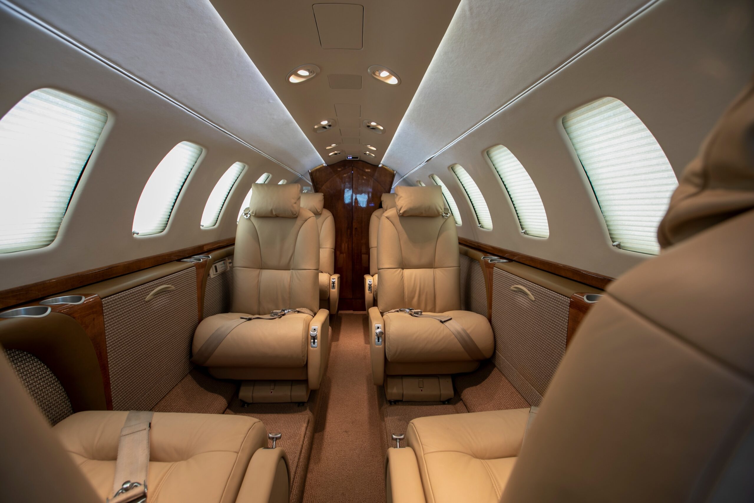 Interior of light jet with beige seats and window blinds down. Looking from front of cabin towards rear