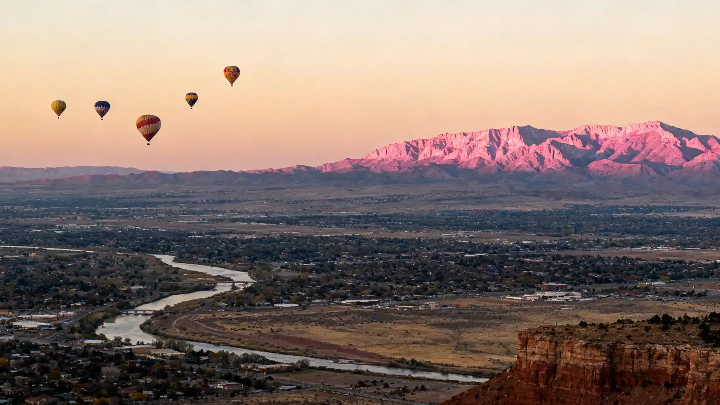 A wide view of the Rio Grande valley at Albuquerque with several hot-air balloons floating far away in the sky and the Sandia Mountains glowing pink on the horizon in golden-hour light
