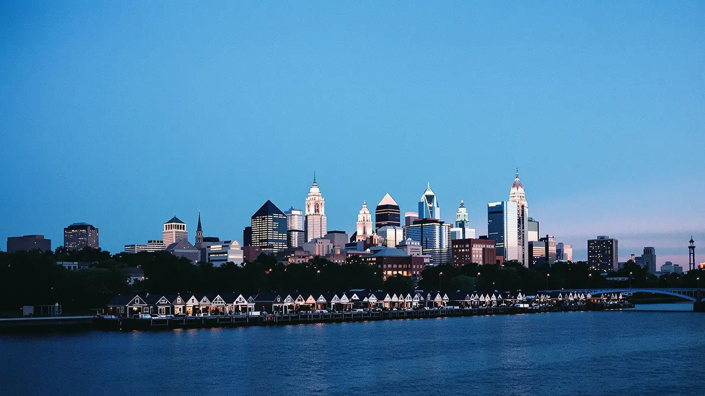 The Philadelphia skyline above the Schuylkill River with a row of historic boathouses along the bank at dusk